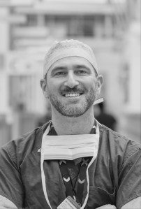 An image of Giangiacomo Ollandini in scrubs and surgical cap, wearing a face mask around his neck
